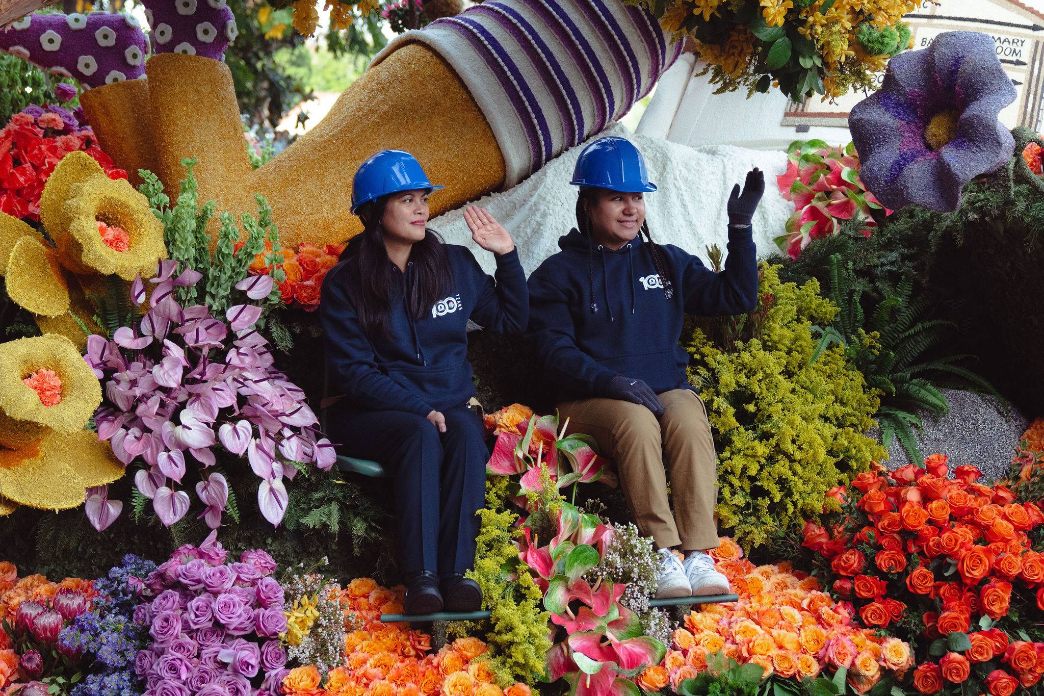 Two people wearing blue hard hats waving from a flower-covered float at the Rose Parade.