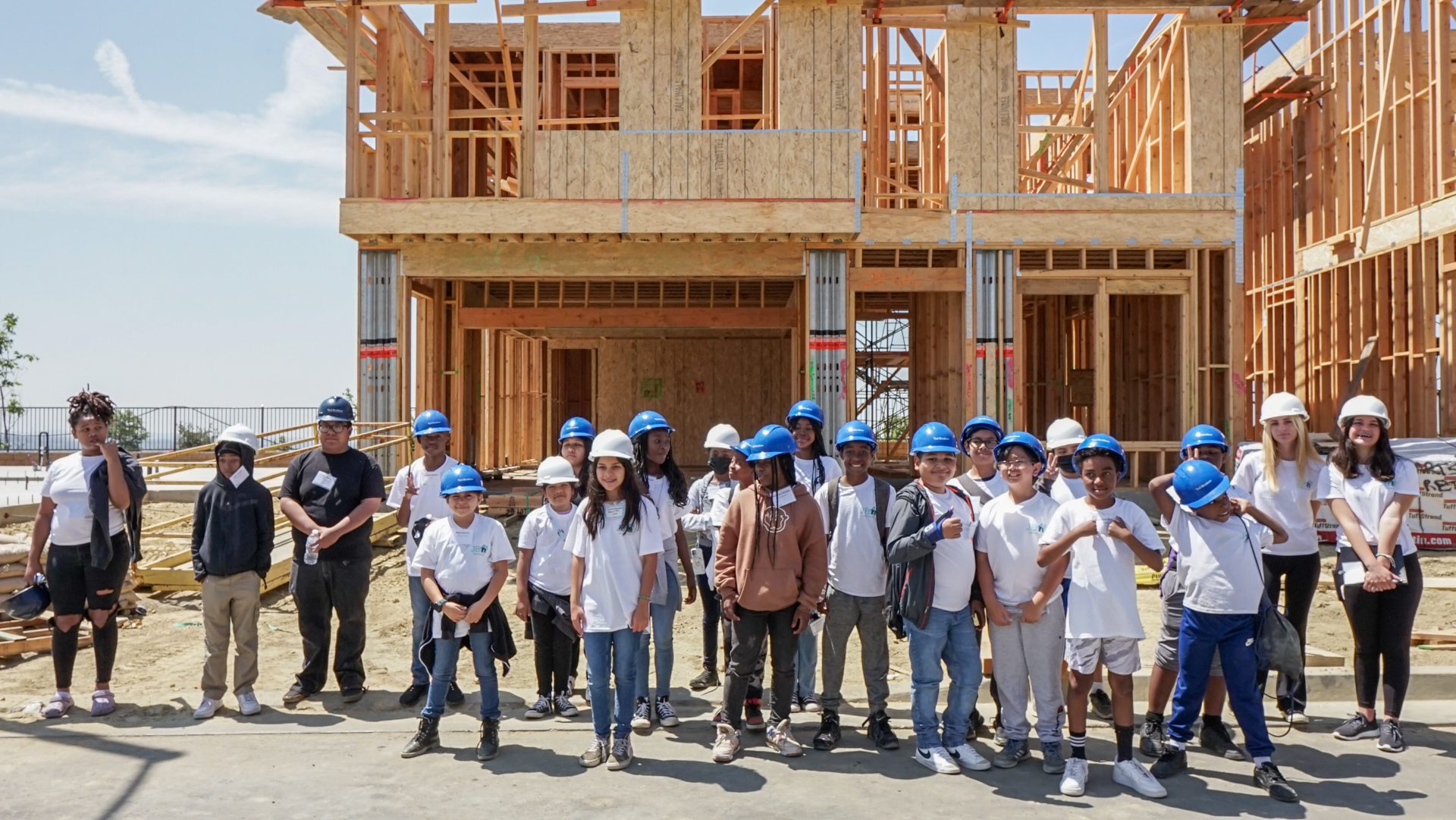 Group of young students in hard hats at a home building education site, smiling and posing for a photo.