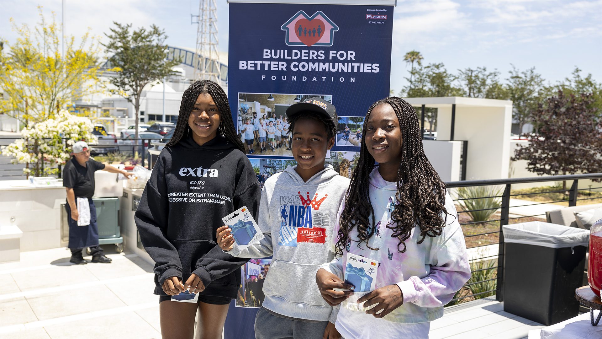 Three young students at a Builders for Better Communities event, holding ID badges and smiling.